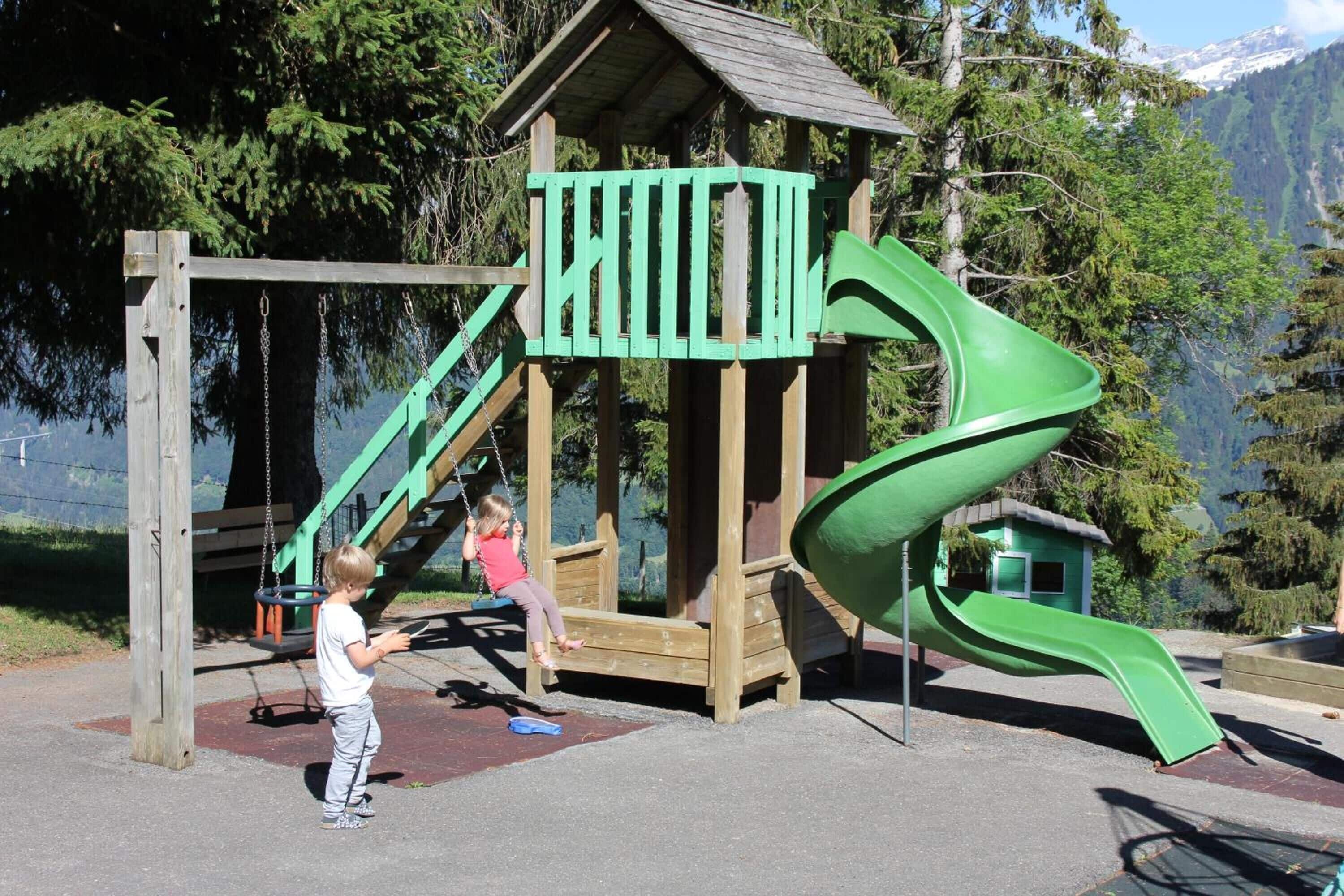 A playground with a wooden playhouse, a green slide, and swings.