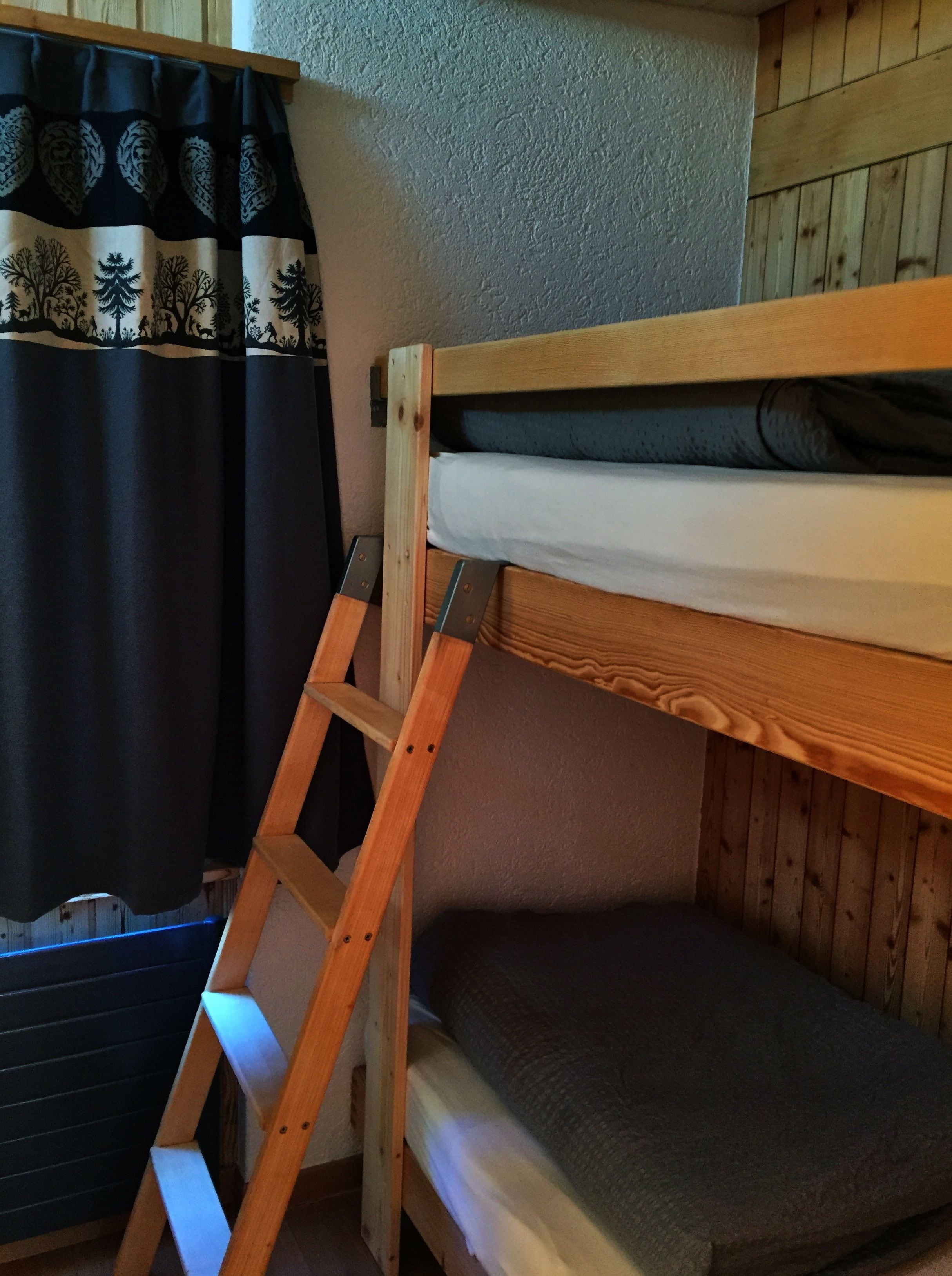 A bunk bed with a wooden ladder, a window with curtains, and a wooden wall.