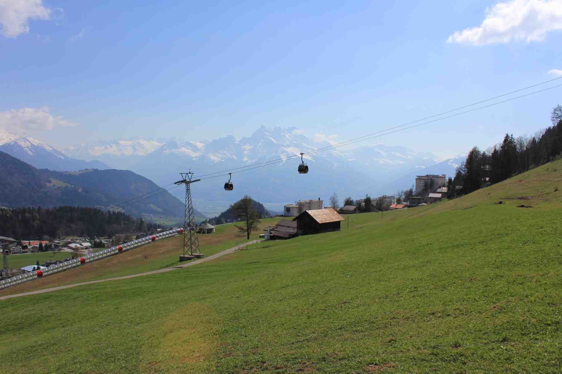 A green hillside with a small building, a cable car, and a mountain range in the background.