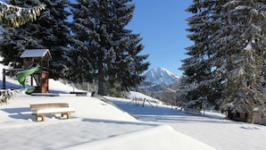 Un parque cubierto de nieve con un banco de madera, un tobogán verde y una montaña al fondo.