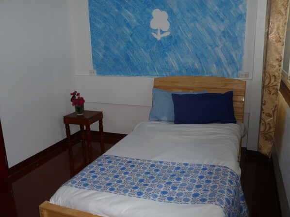 Une chambre avec un lit en bois, une couvre-lit à motifs floraux bleus, une petite table de chevet en bois ornée d’un vase de fleurs et un tableau abstrait bleu accroché au mur.