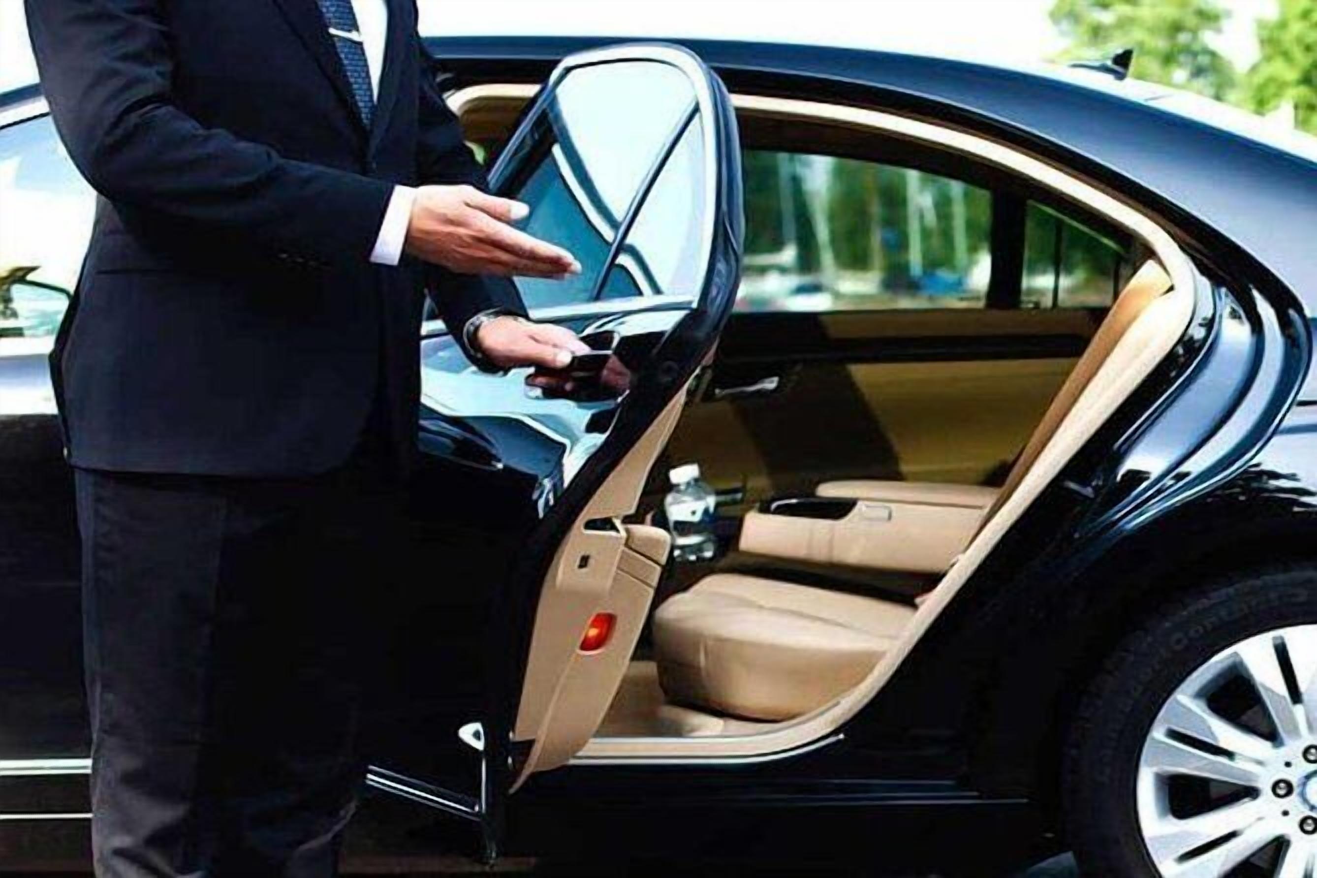 A person in a suit standing next to a car with an open door, revealing the interior.