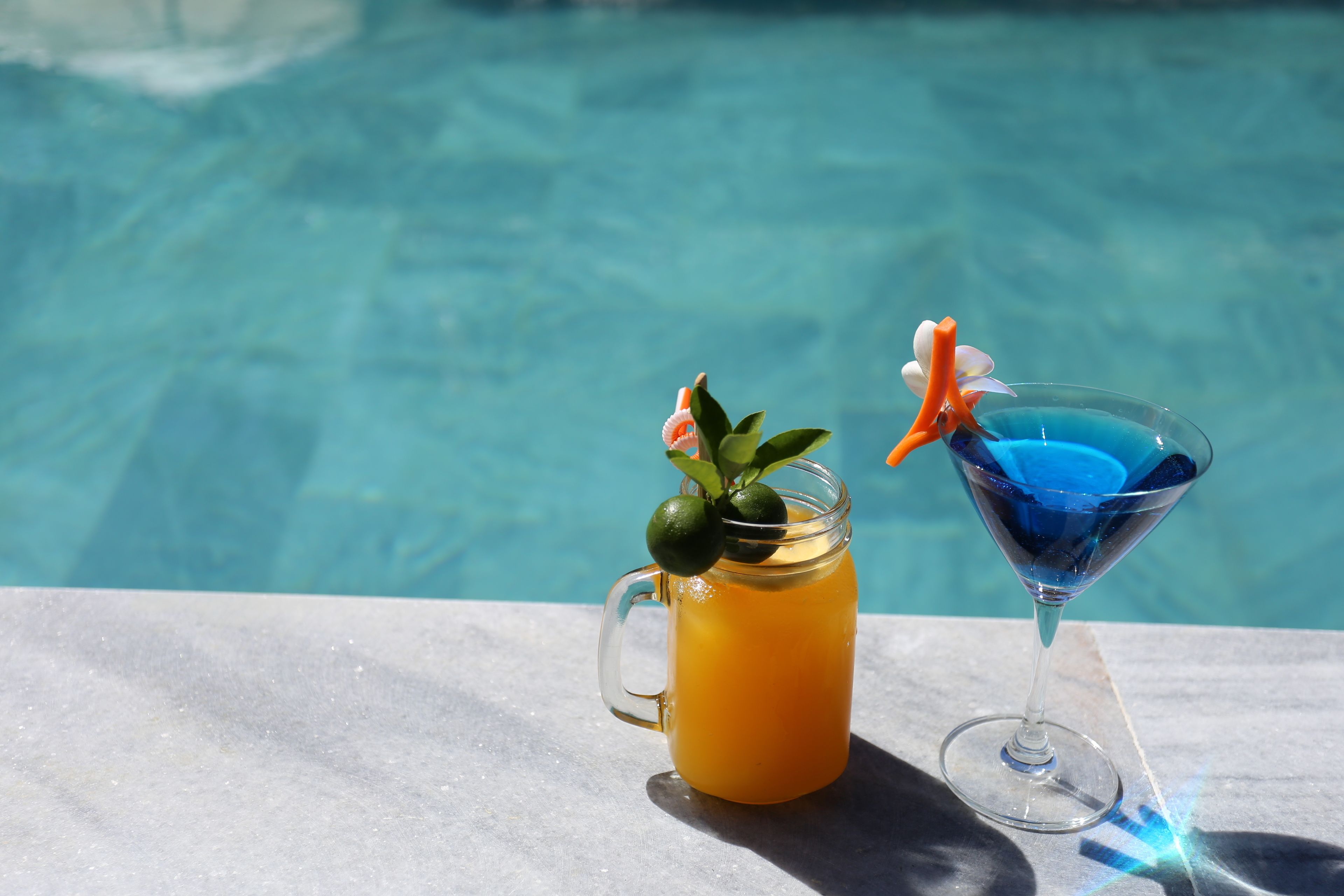 A glass of orange juice and a martini glass by a poolside.