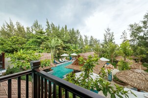 Área de piscina en un resort con sombrillas de paja, sillones de descanso y abundante vegetación.