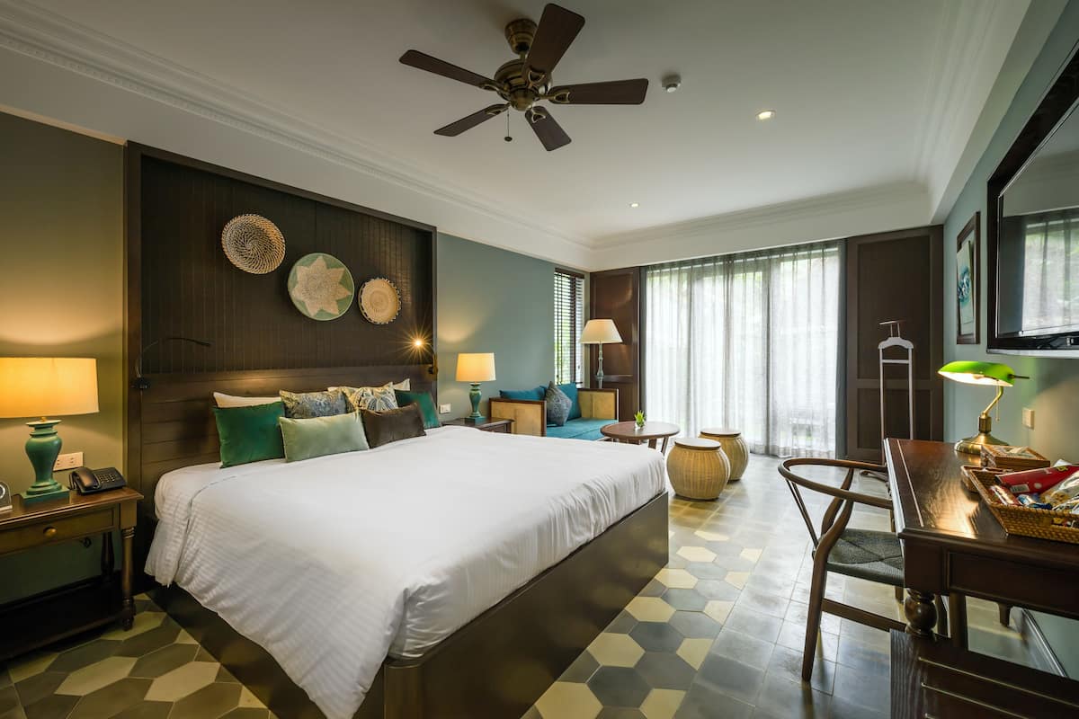 A hotel room with a large bed, a desk, a chair, a ceiling fan, and a TV.