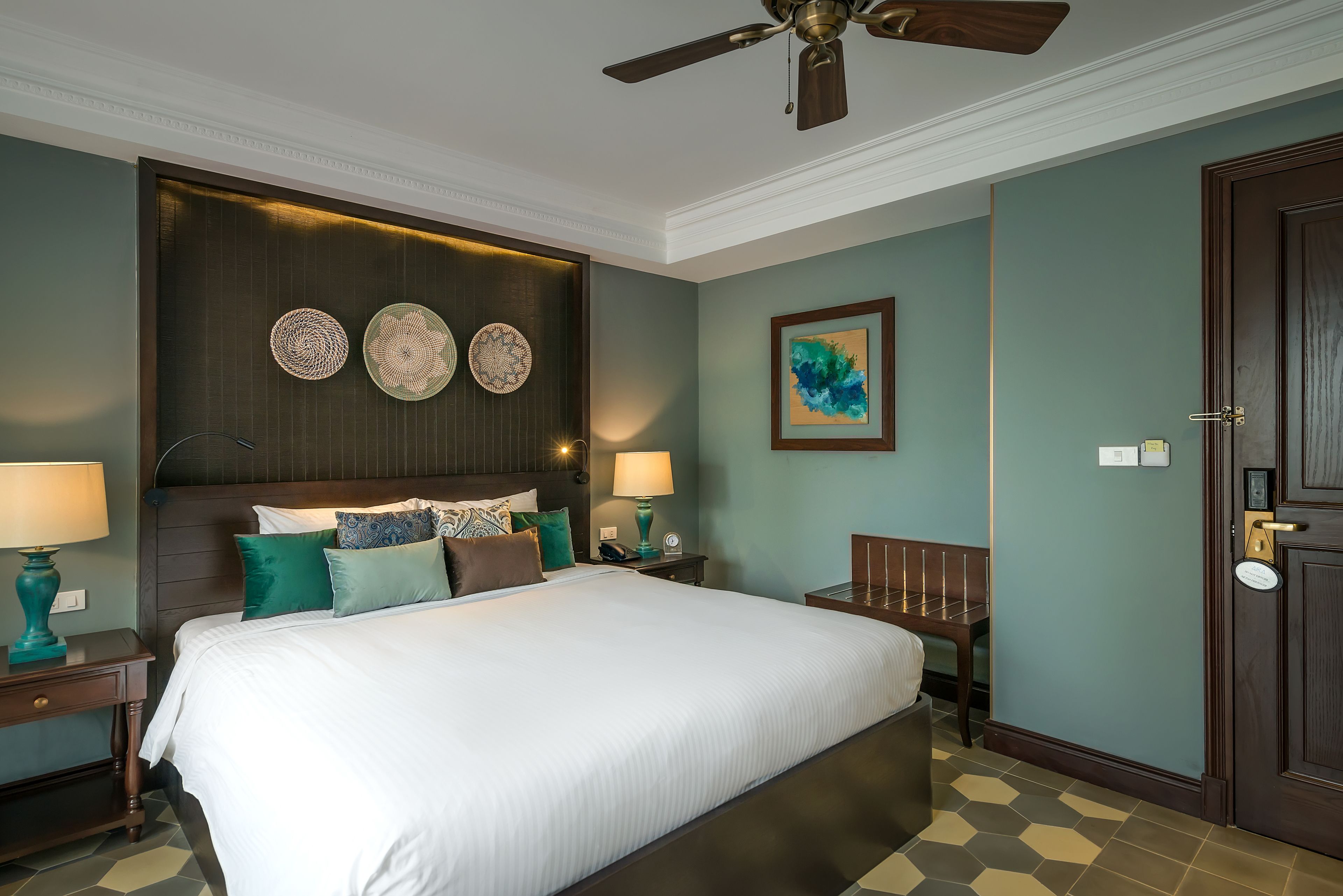 A neatly arranged hotel room with a large bed, two bedside tables with lamps, a ceiling fan, and a door with a keypad lock.