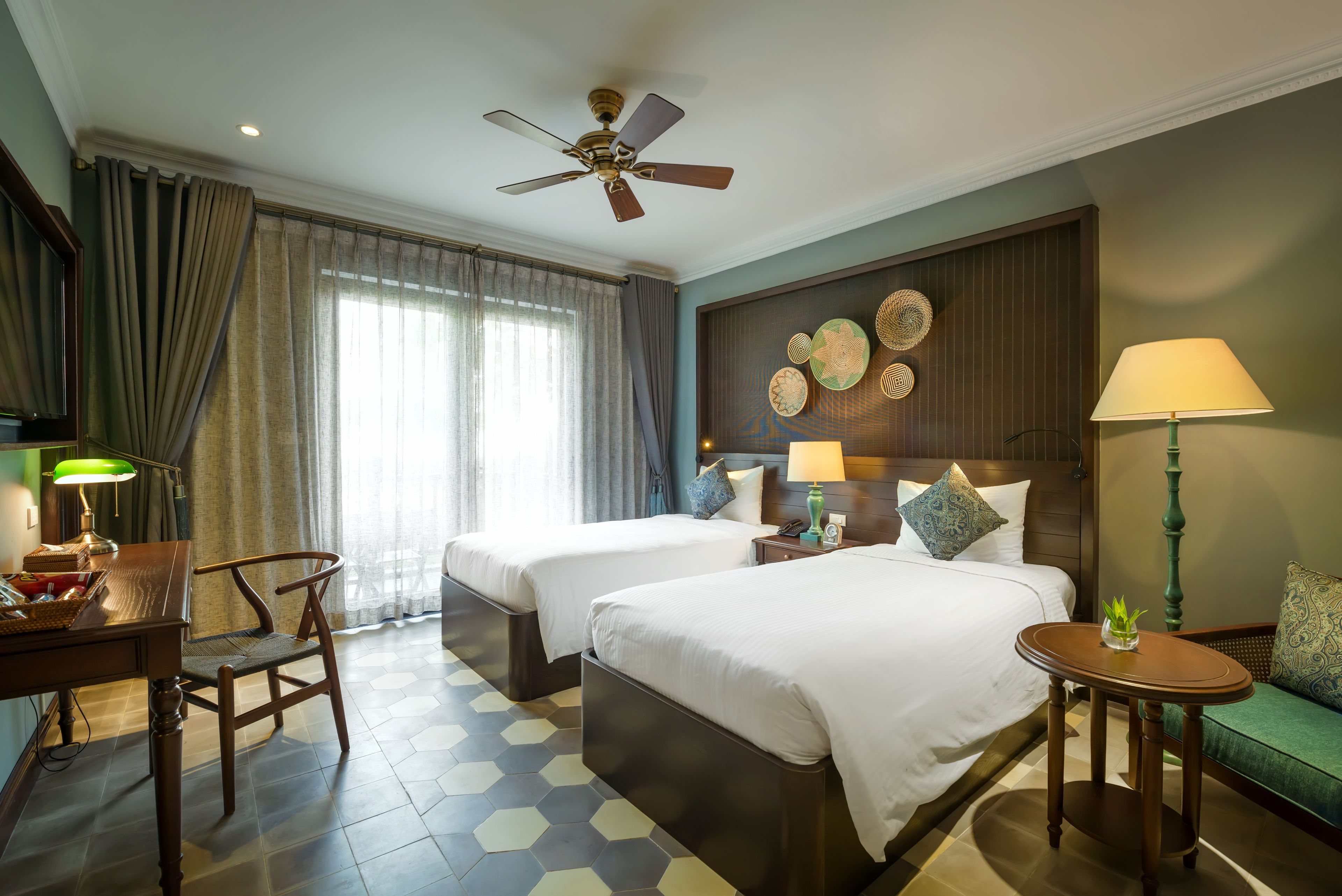 A hotel room with two beds, a desk, a chair, a ceiling fan, and a green sofa.