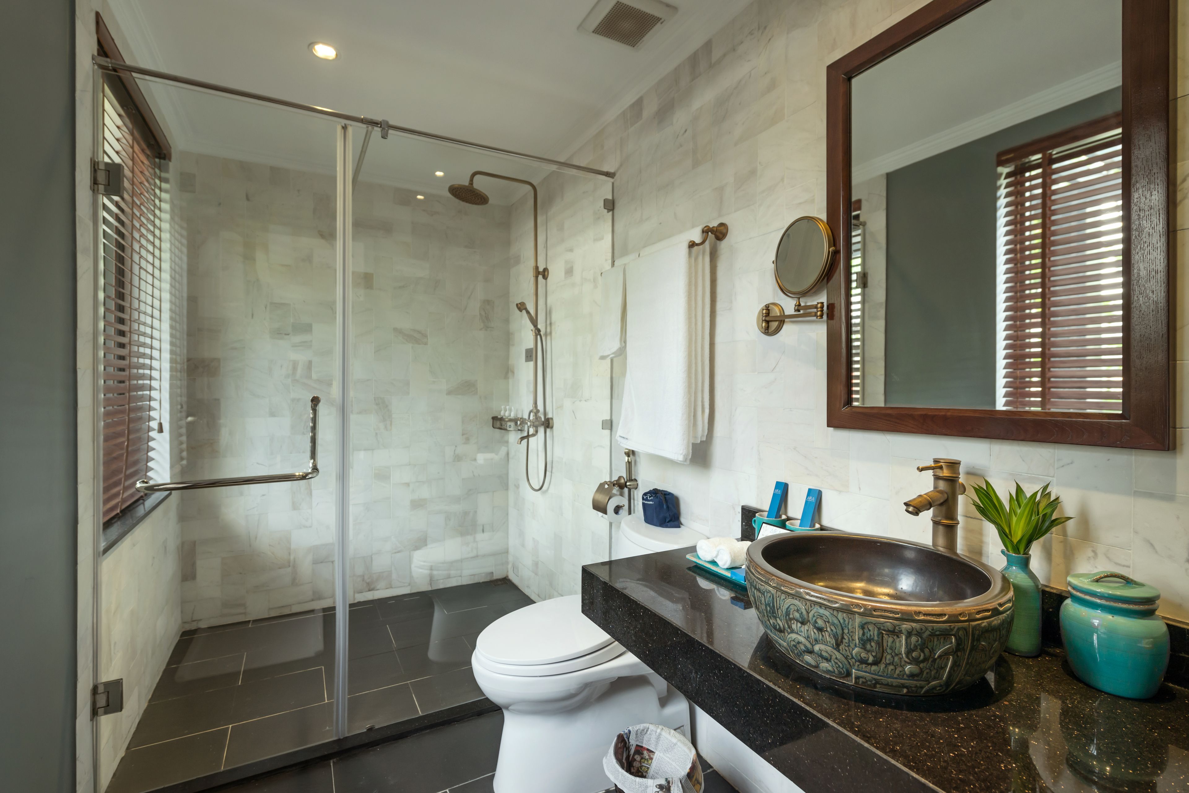 Un baño moderno con cabina de ducha de vidrio, inodoro blanco, espejo grande y encimera de granito con un lavabo decorativo.