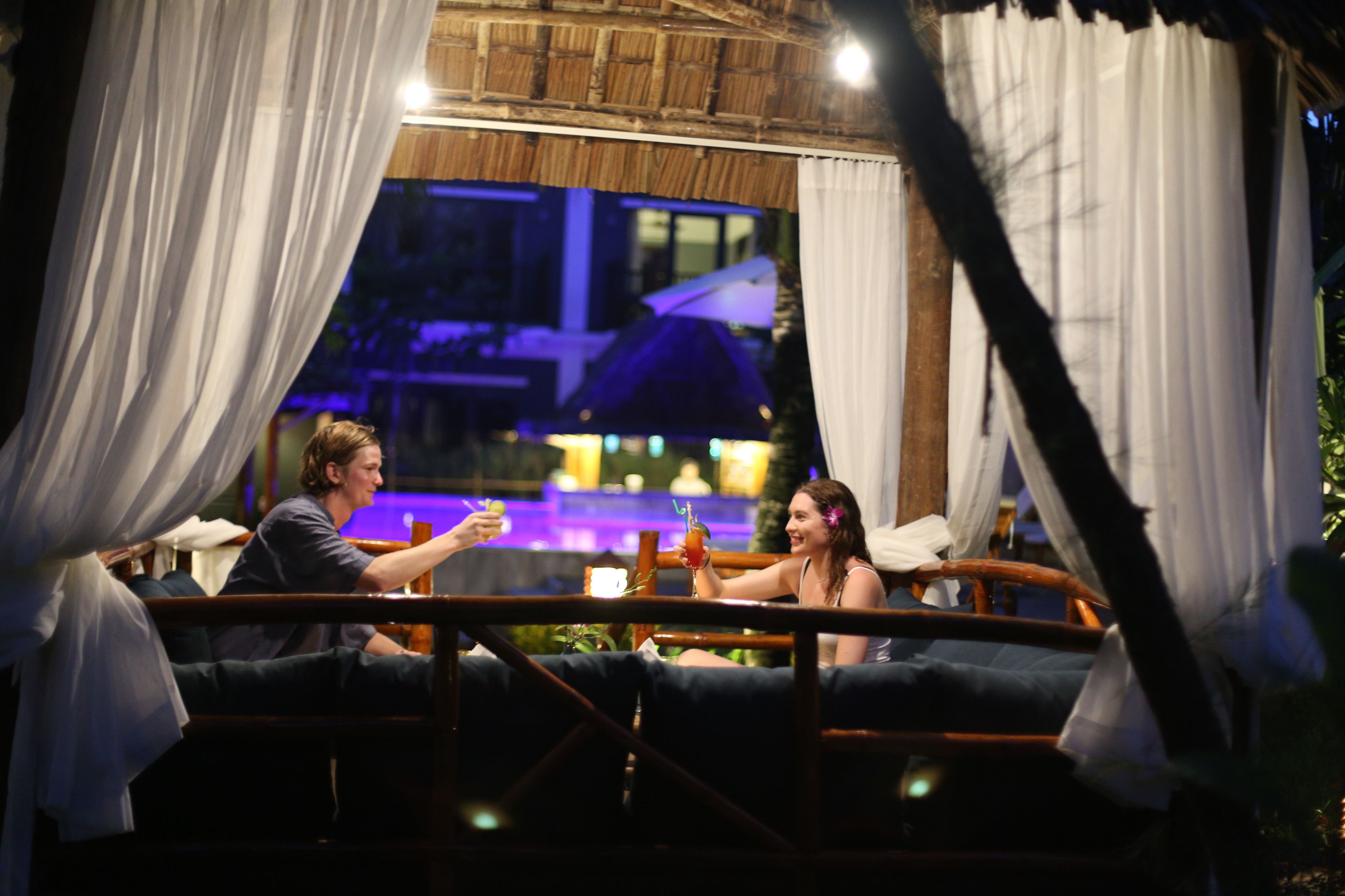 Una pareja sentada en un banco de madera con cortinas blancas, disfrutando de bebidas en un ambiente exterior con poca luz.