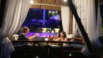 Una pareja sentada en un banco de madera con cortinas blancas, disfrutando de bebidas en un ambiente exterior con poca luz.