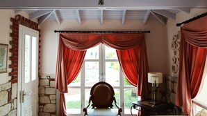 Una habitación con techo alto, vigas a la vista, candelabro, cortinas rojas, pared de piedra, banco de madera y una mesa con lámpara.