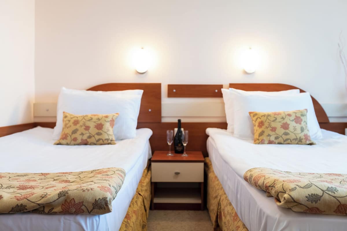 Habitación de hotel con dos camas, cada una con ropa de cama y almohadas estampadas con motivos florales, una mesita de noche con una botella y vasos, y luces fijadas a la pared.
