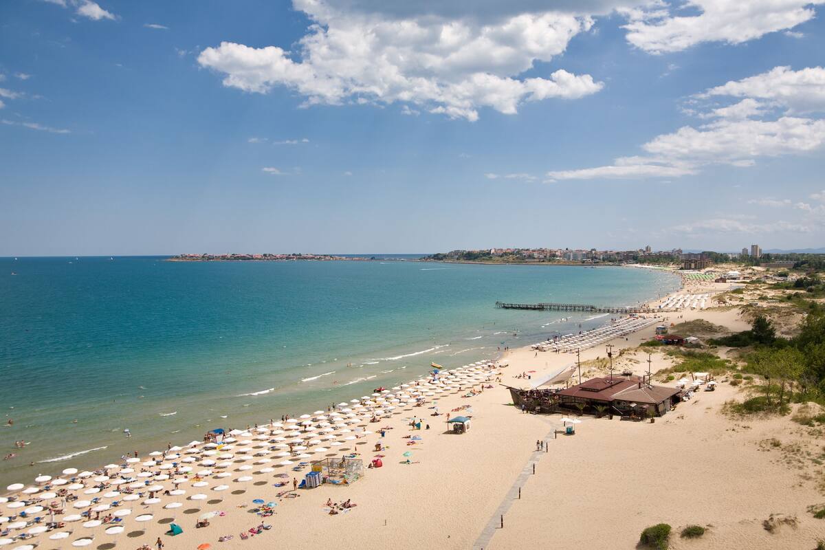 Una playa con numerosos sombrillas y tumbonas, un muelle que se adentra en el agua y un paisaje urbano al fondo.