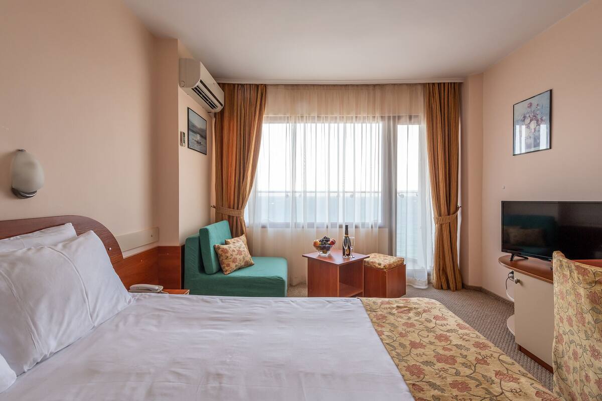 Habitación de hotel con una cama grande, un sofá verde, una mesita con una botella y fruta, un televisor y una ventana con cortinas.