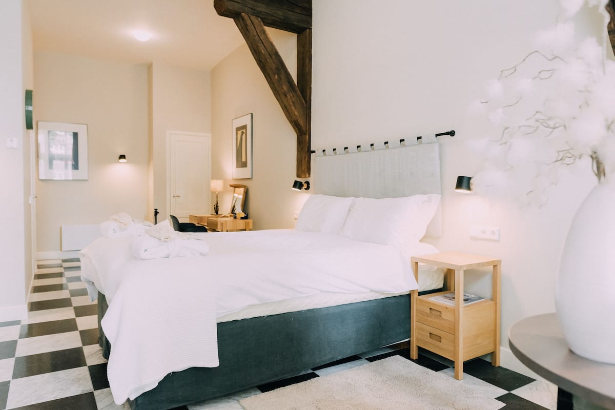 Un dormitorio con piso a cuadros, una cama con ropa blanca, una mesita de noche de madera y una lámpara en la mesita junto a la cama.