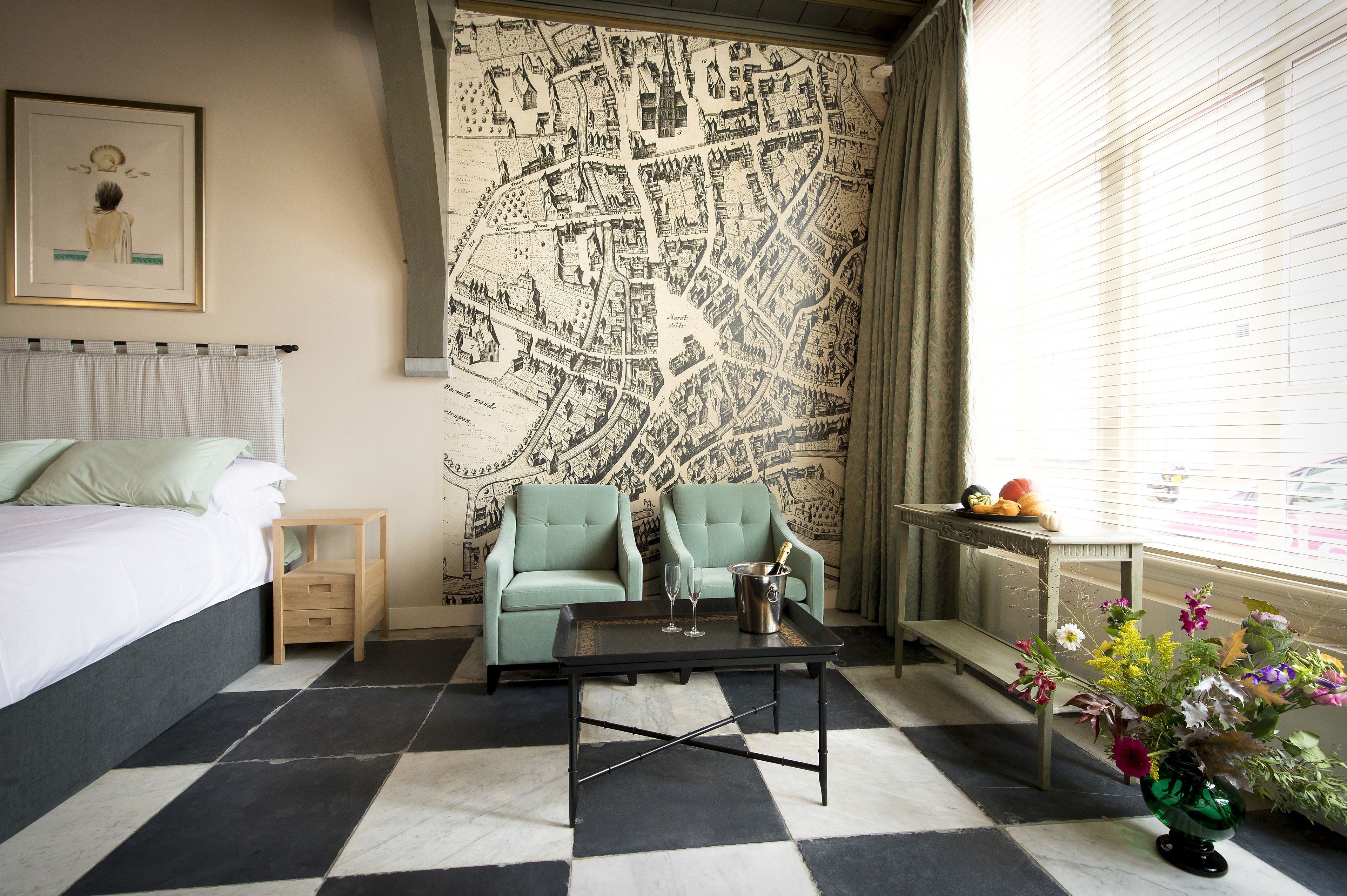 Hotelkamer met een bed, twee fauteuils, een bijzettafeltje, een kaart aan de muur, een raam met zonwering en een vaas met bloemen.