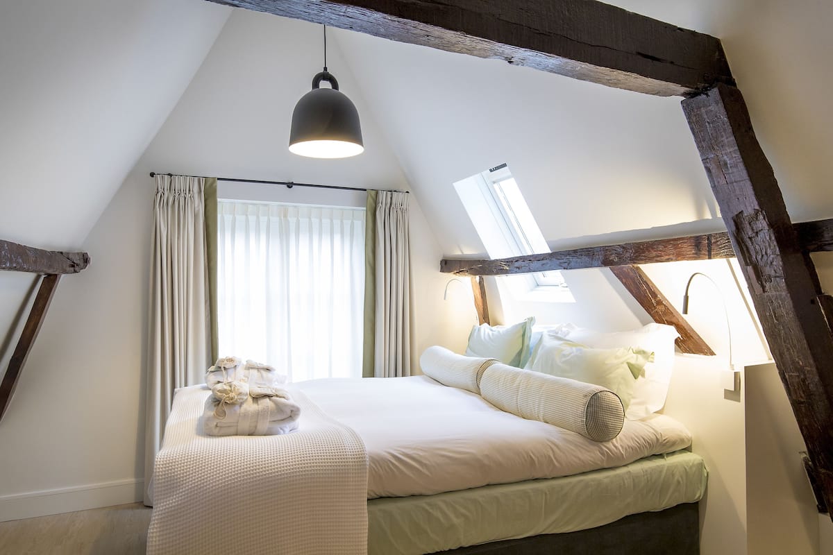 Un dormitorio con techo inclinado, una cama con almohadas y una manta, un lámpara colgante y una ventana con cortinas.