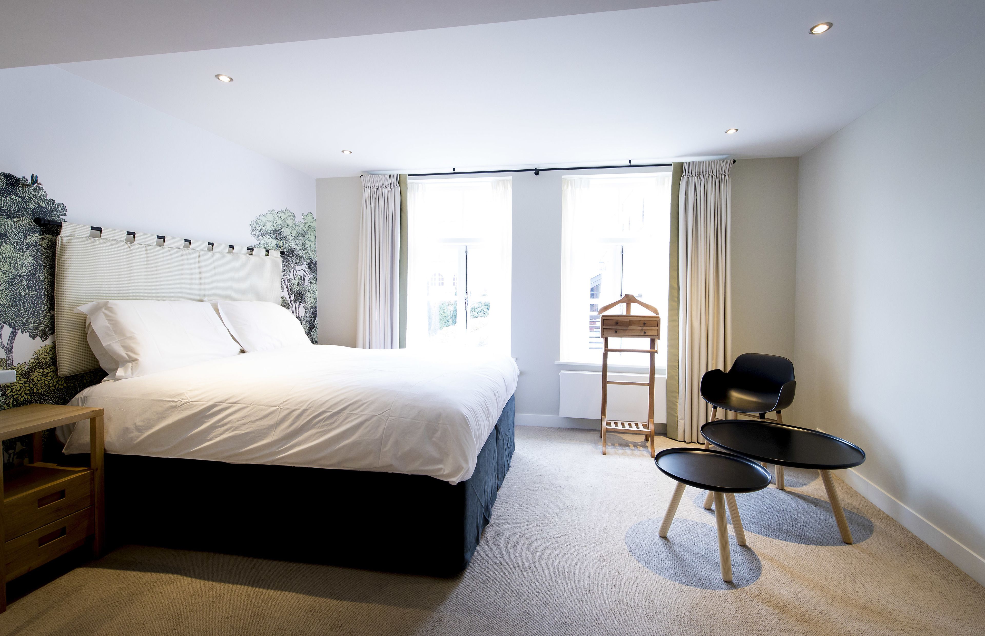 A modern bedroom with a large bed, a wooden nightstand, a small table, two chairs, and a window with curtains.
