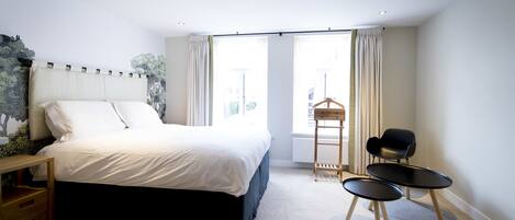 A modern bedroom with a large bed, a wooden nightstand, a small table, two chairs, and a window with curtains.