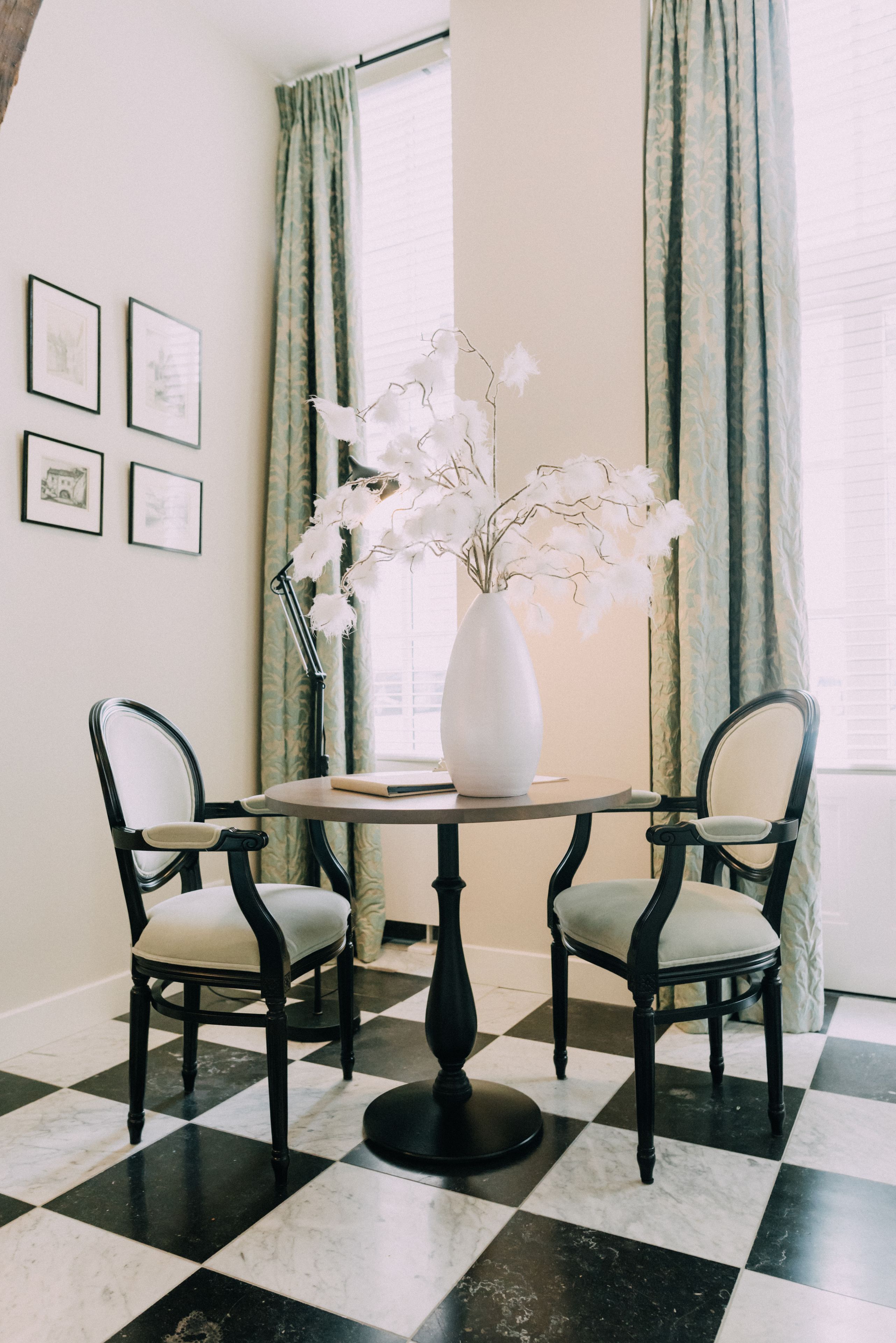 A dining area with a checkered floor, two chairs, a round table, a white vase with flowers, and curtains.