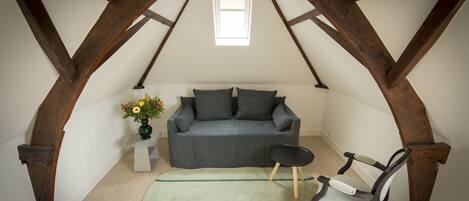 A loft bedroom with exposed wooden beams, a grey sofa, a small round table, and a green rug.