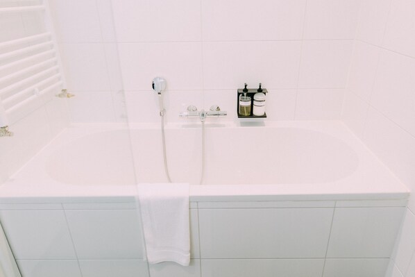 Une baignoire blanche, avec un pommeau de douche à main, une serviette et deux bouteilles de shampoing posées sur le bord.