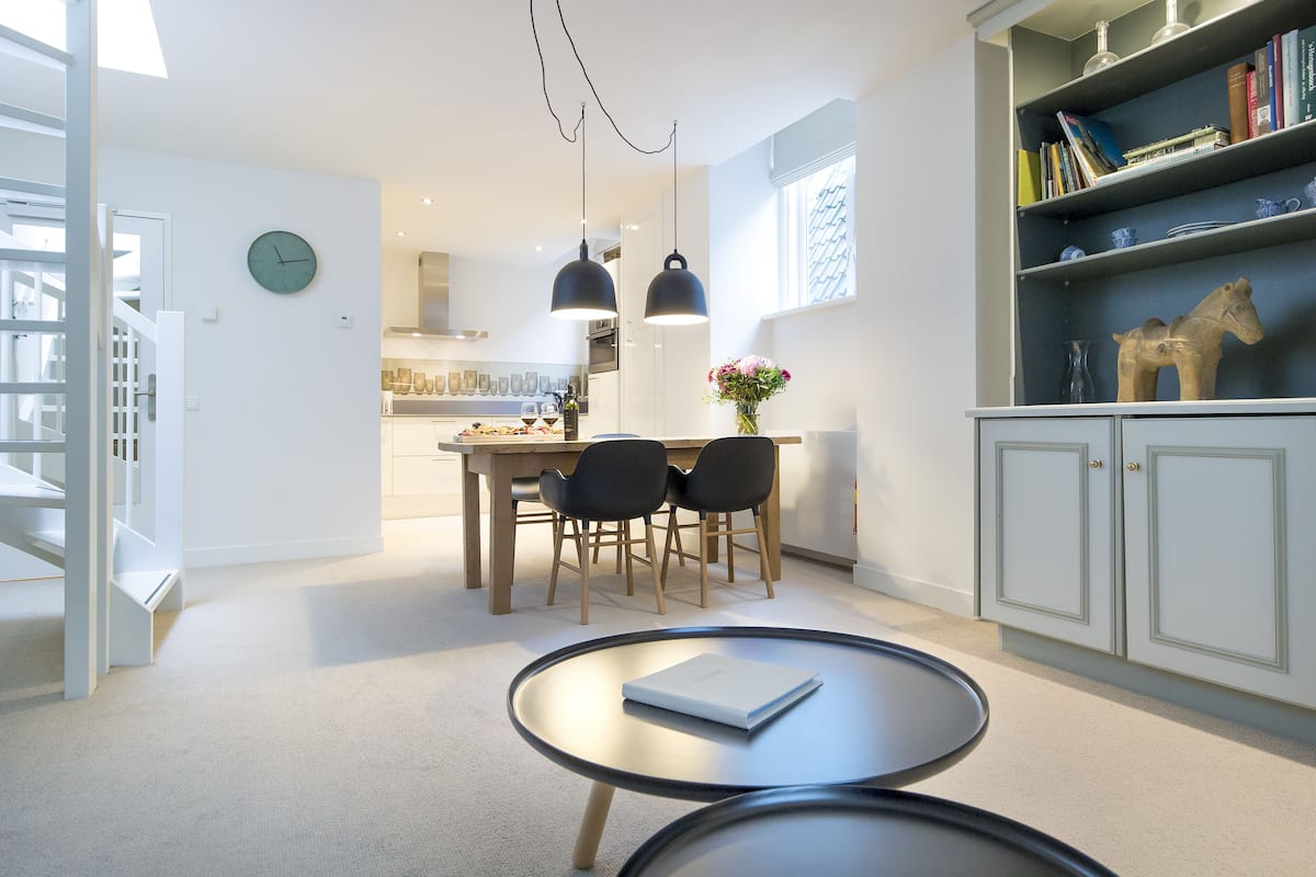 Una cocina moderna con luces colgantes, un comedor con mesa y sillas, y una estantería con libros y objetos decorativos.