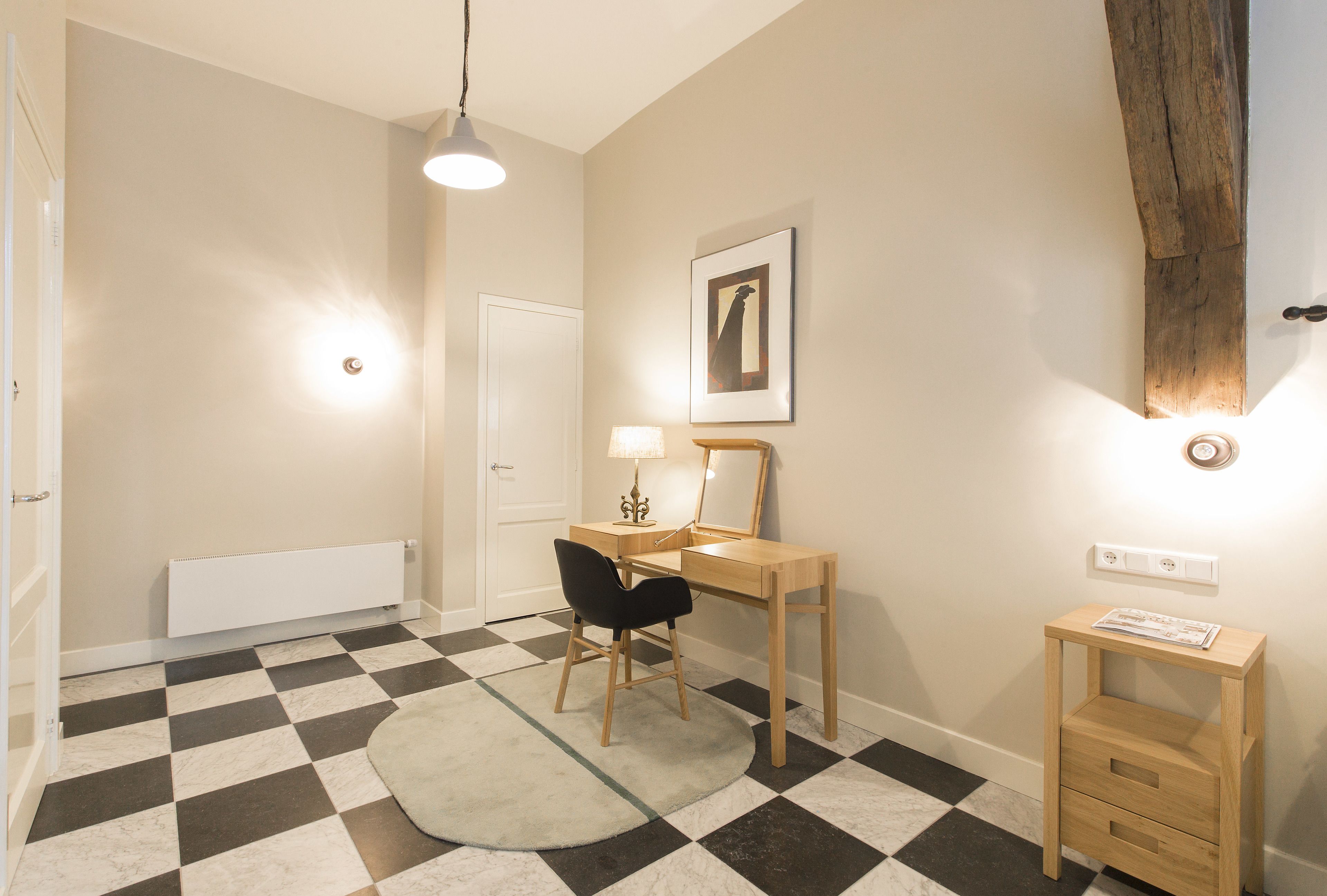 Una habitación con piso a cuadros, un escritorio con una silla, una lámpara, un espejo y una pequeña mesita de madera.