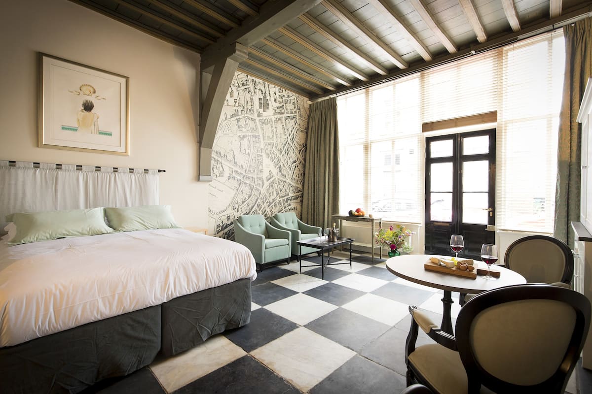 Habitación de hotel con cama, dos sillones, una mesita con una botella y vasos, y una ventana con cortinas.