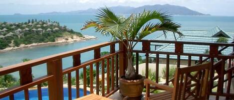 A balcony with a wooden table and chairs, overlooking a swimming pool and a coastal view with houses and mountains in the distance.