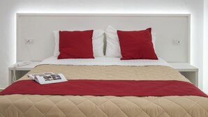 Una cama con una colcha beige acolchada y almohadas rojas, una mesita de noche blanca con un teléfono y una revista.
