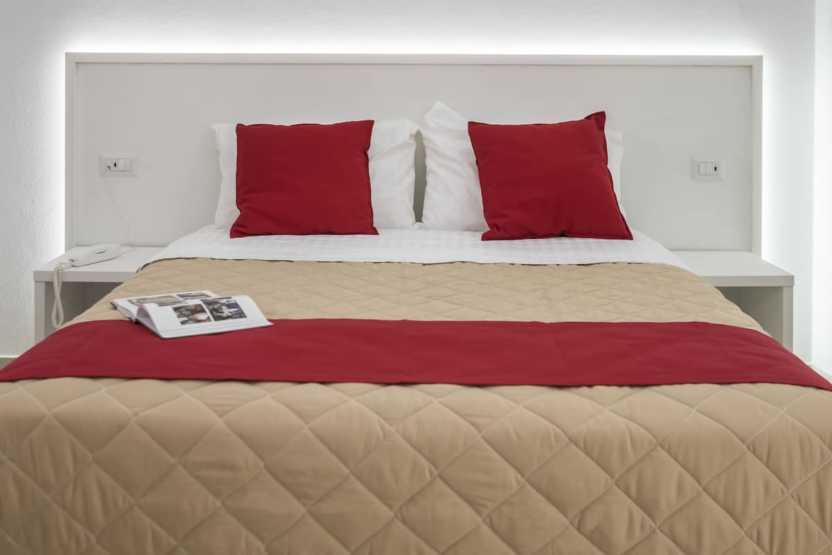 Uma cama com colcha bege acolchoada e almofadas vermelhas, uma mesa de cabeceira branca com um telefone e uma revista.