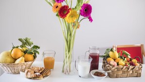 Una mesa de desayuno con diversas frutas, un jarrón de flores y dos jarras de bebidas.