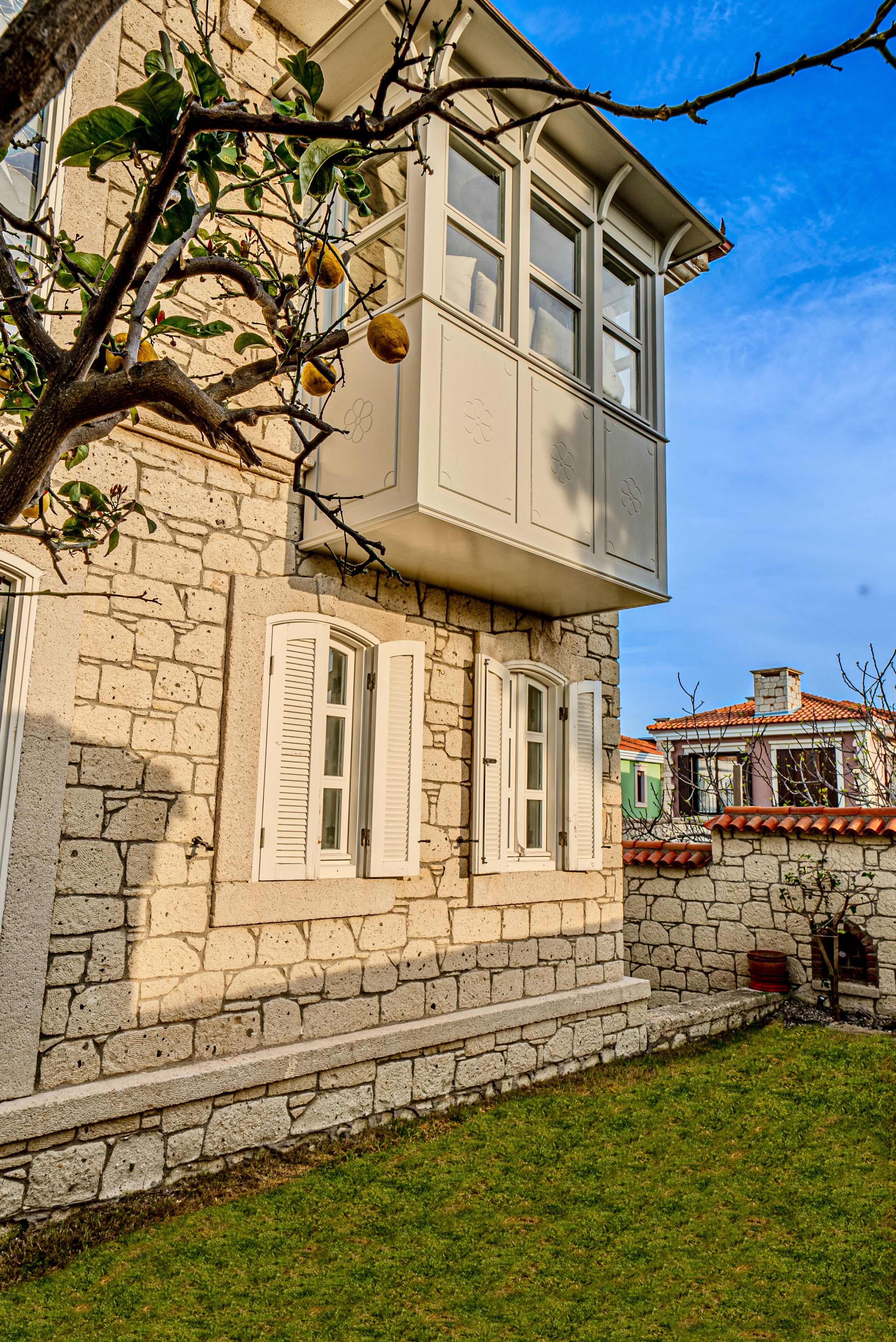 Um edifício de pedra com varanda, janelas com persianas e uma árvore com frutos.