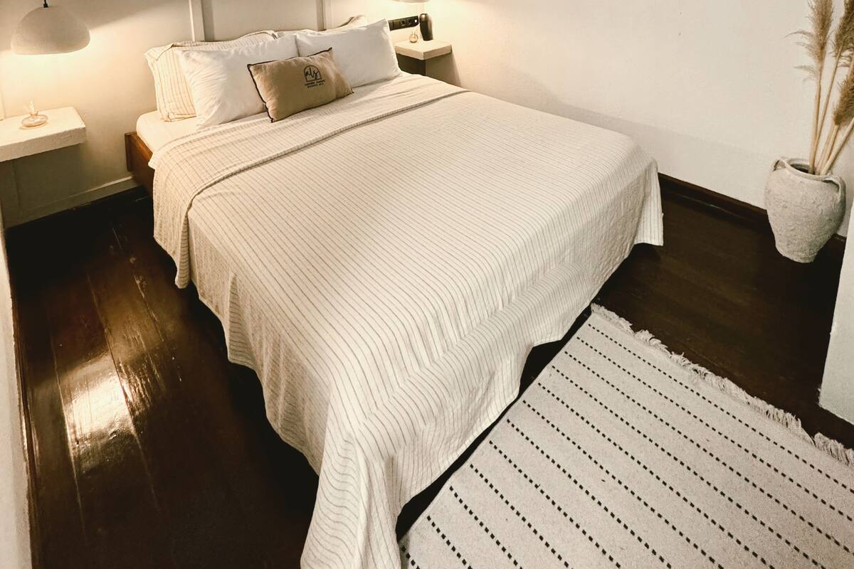 Un dormitorio con una cama grande, una mesita de noche, una lámpara, un candelabro colgante y una ventana con persianas.