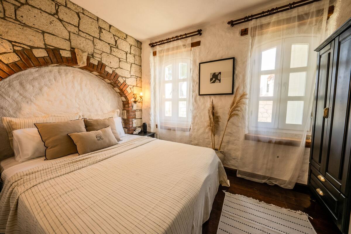 Un dormitorio con pared de piedra, una cama grande con cabecera, un armario y una ventana con cortinas.