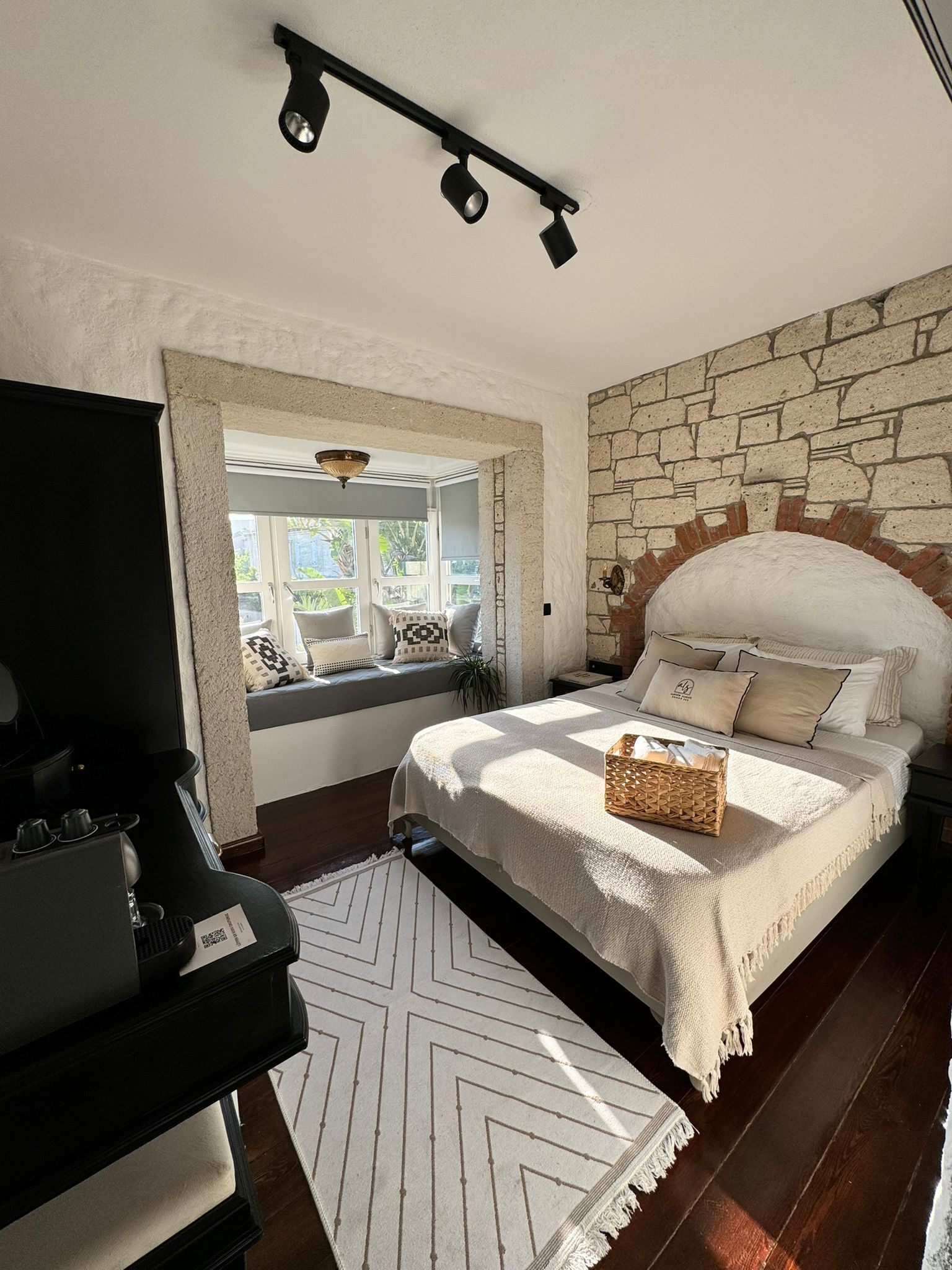 A bedroom with a stone wall, a bed with a white headboard, a window seat with cushions, a black TV stand, and a woven basket on the bed.