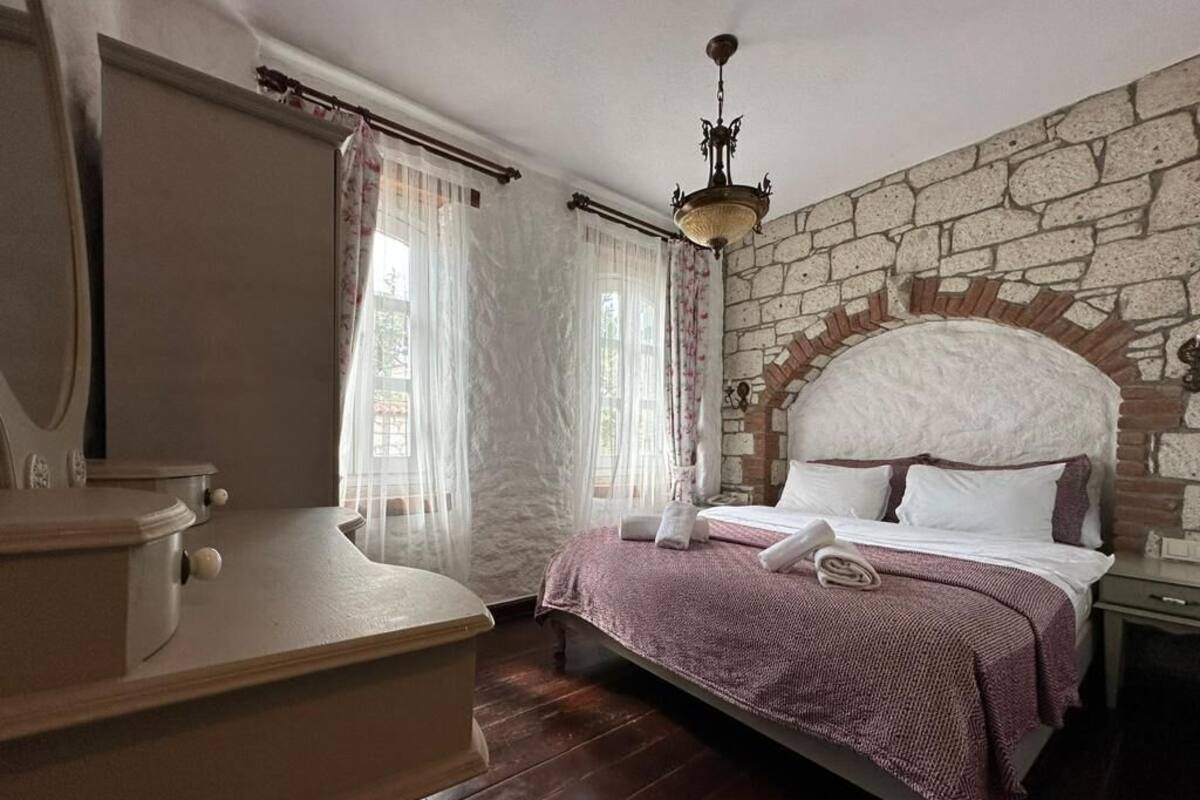 Un dormitorio con pared de piedra, una cama con cabecera, una mesita de noche, un aparador y una araña.