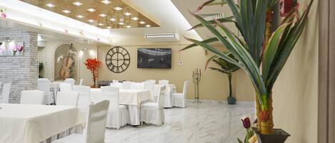 A spacious dining area with white tables and chairs, a large wall clock, and a modern ceiling design.