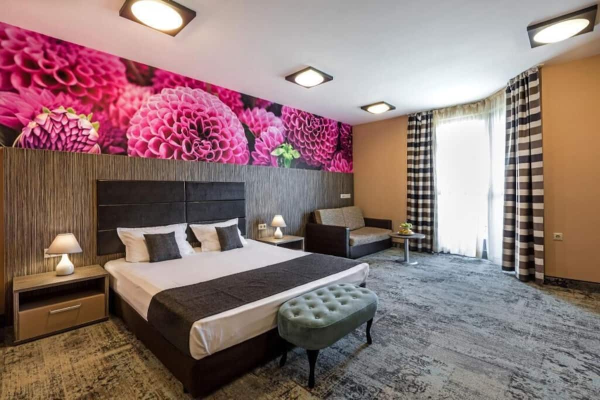 A hotel room with a large bed, two bedside tables, a bench, a wall mural, and a window with curtains.