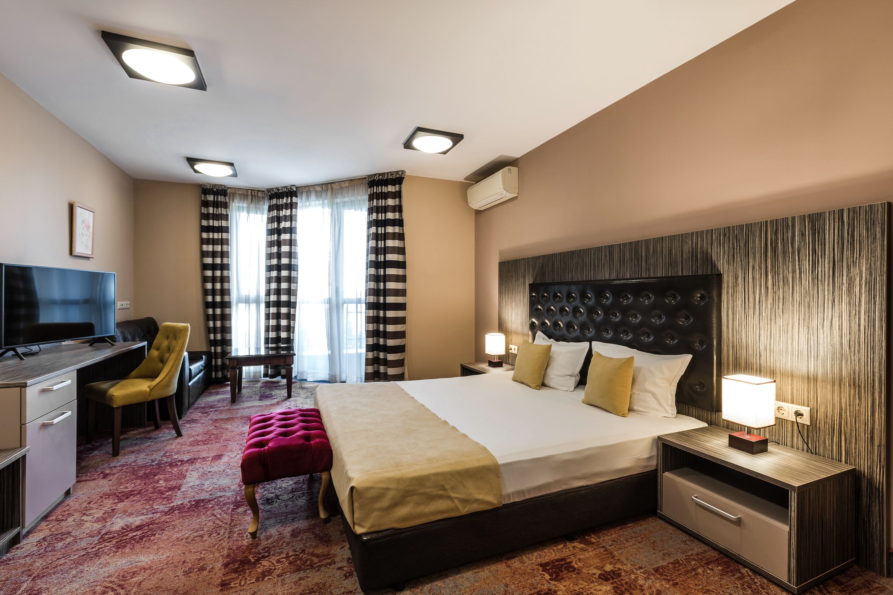 A hotel room with a large bed, a desk with a chair, a television, and a red ottoman.