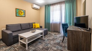 A modern living room with a grey sofa, a white coffee table, a blue armchair, and a TV on a wooden stand. - Plaza Hotel (Plovdiv)