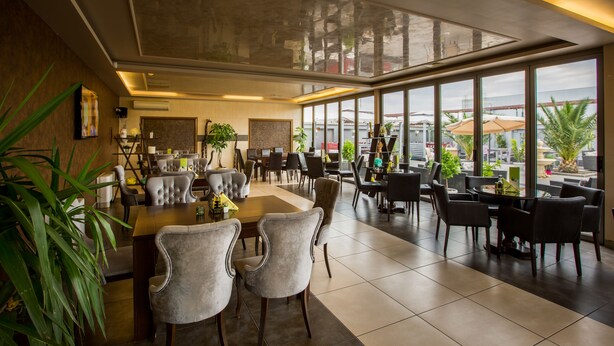 A spacious dining area with a long table and multiple chairs, large windows offering a view of the outdoors, and potted plants adding greenery. - Plaza Hotel (Plovdiv)