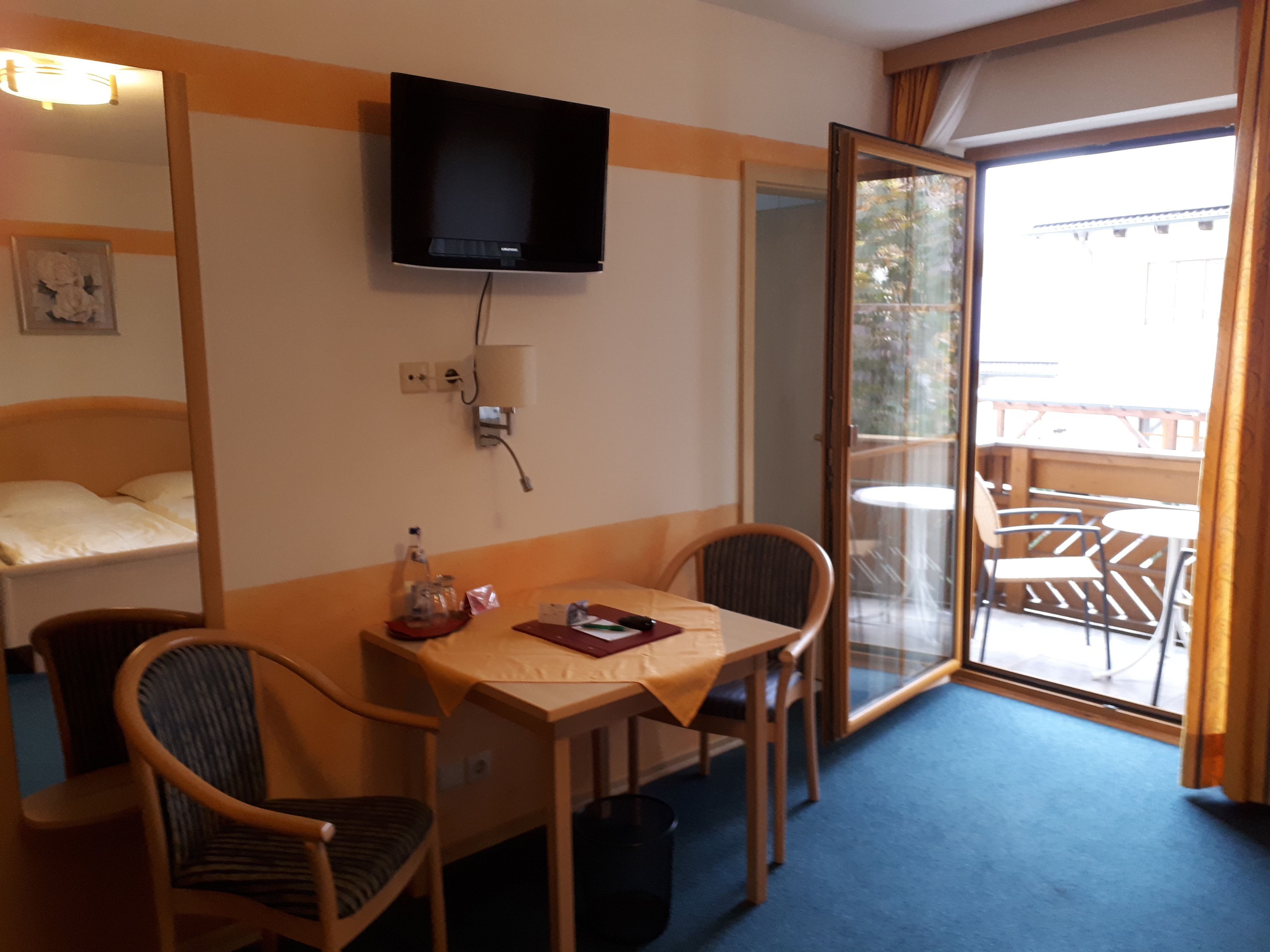 Habitación de hotel con cama, televisión montada en la pared, una mesa con sillas y una puerta corrediza de vidrio que da a un espacio exterior con mesa y sillas.