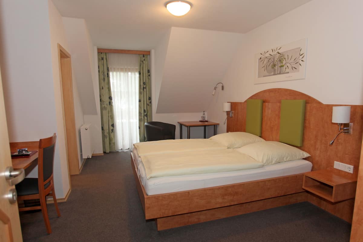 A hotel room with a bed, a desk, a chair, a small table, and a lamp.