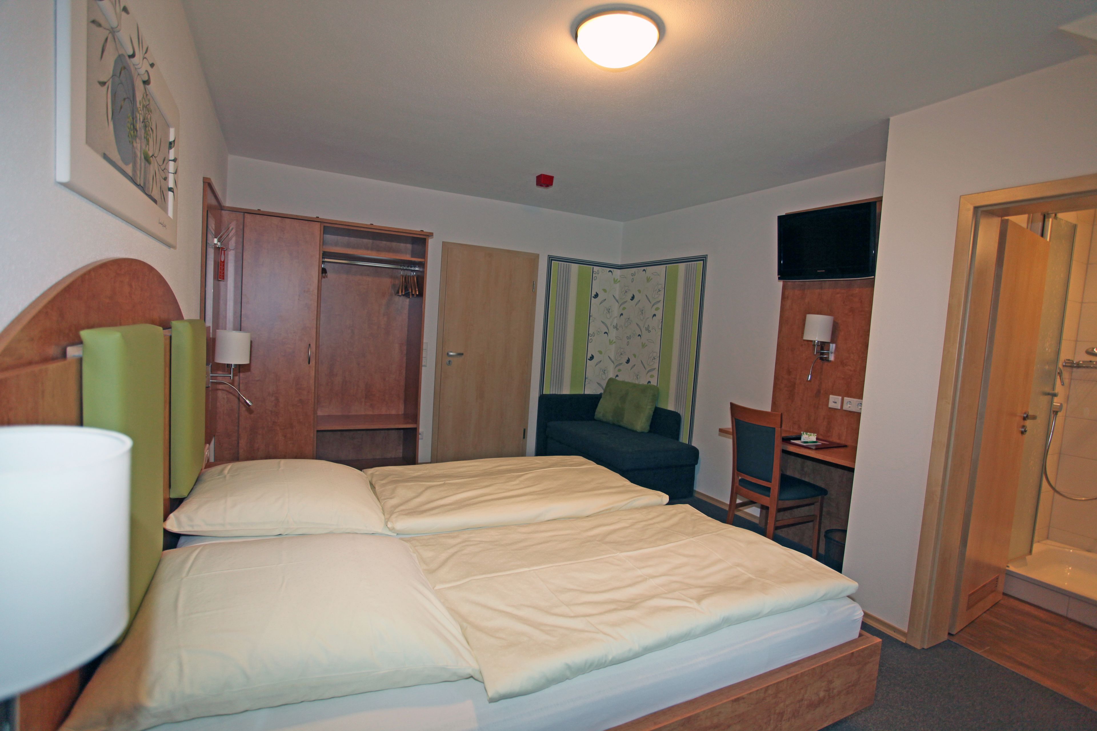 A hotel room with a bed, a desk, a chair, a TV, and a shower.