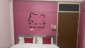Un llit ben fet amb roba de llit blanca i almoinades vermelles, una paret rosa amb un adhesiu de Hello Kitty i una porta tancada.