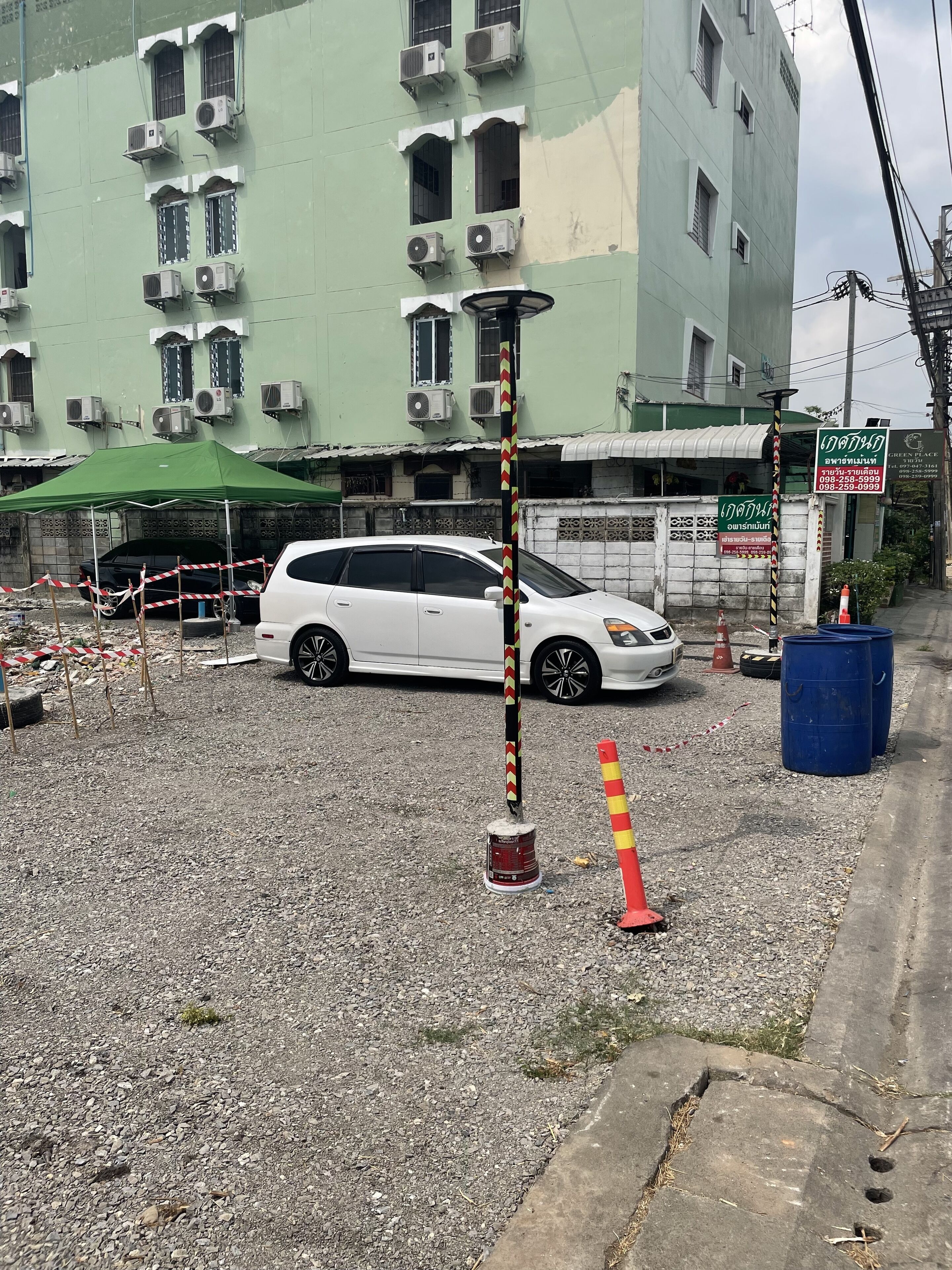 Uma carrinha branca estacionada em frente a um edifício com vários aparelhos de ar condicionado, um toldo verde e uma placa com texto em tailandês.