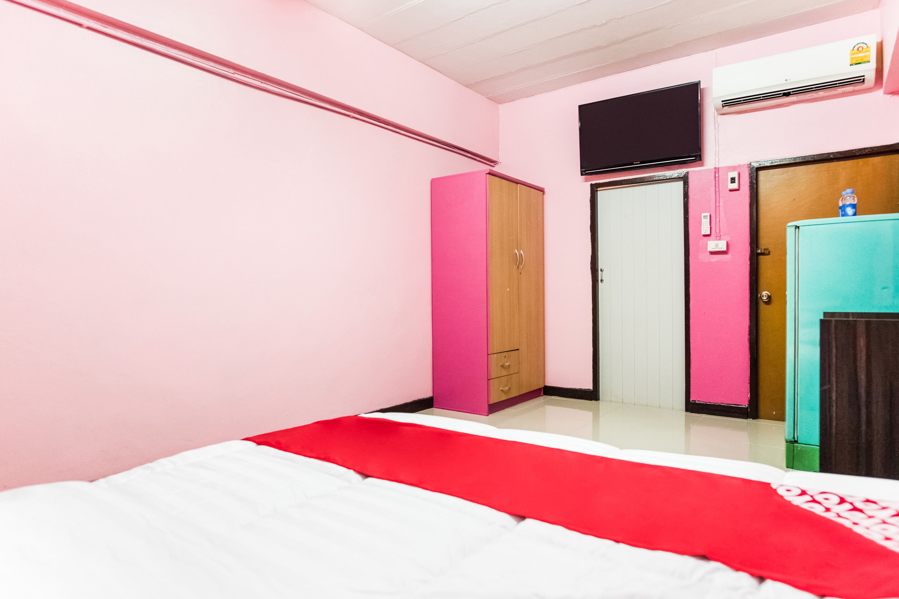 A bedroom with a bed, a pink wardrobe, a teal cabinet, and a television on the wall.