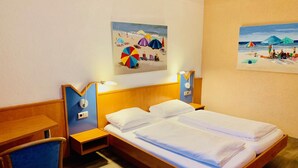 Une chambre d’hôtel avec un lit double, une table de chevet en bois, un bureau et une chaise. On y trouve deux lampes fixées au mur et deux tableaux représentant des scènes de plage.